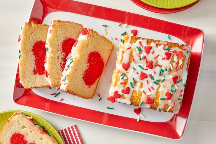 A rectangular loaf cake with white icing and red and green sprinkles sits on a red-edged platter. Three slices reveal a red heart shape inside each piece. Nearby are colorful plates and a striped napkin.