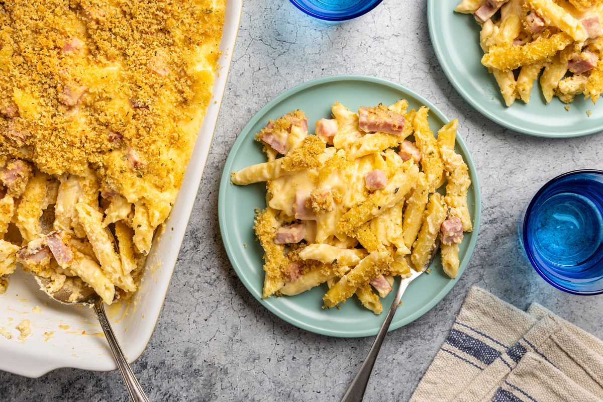 Overhead shot; baked ham pasta with breadcrumb topping on blue plates; water glasses; striped napkin; gray background