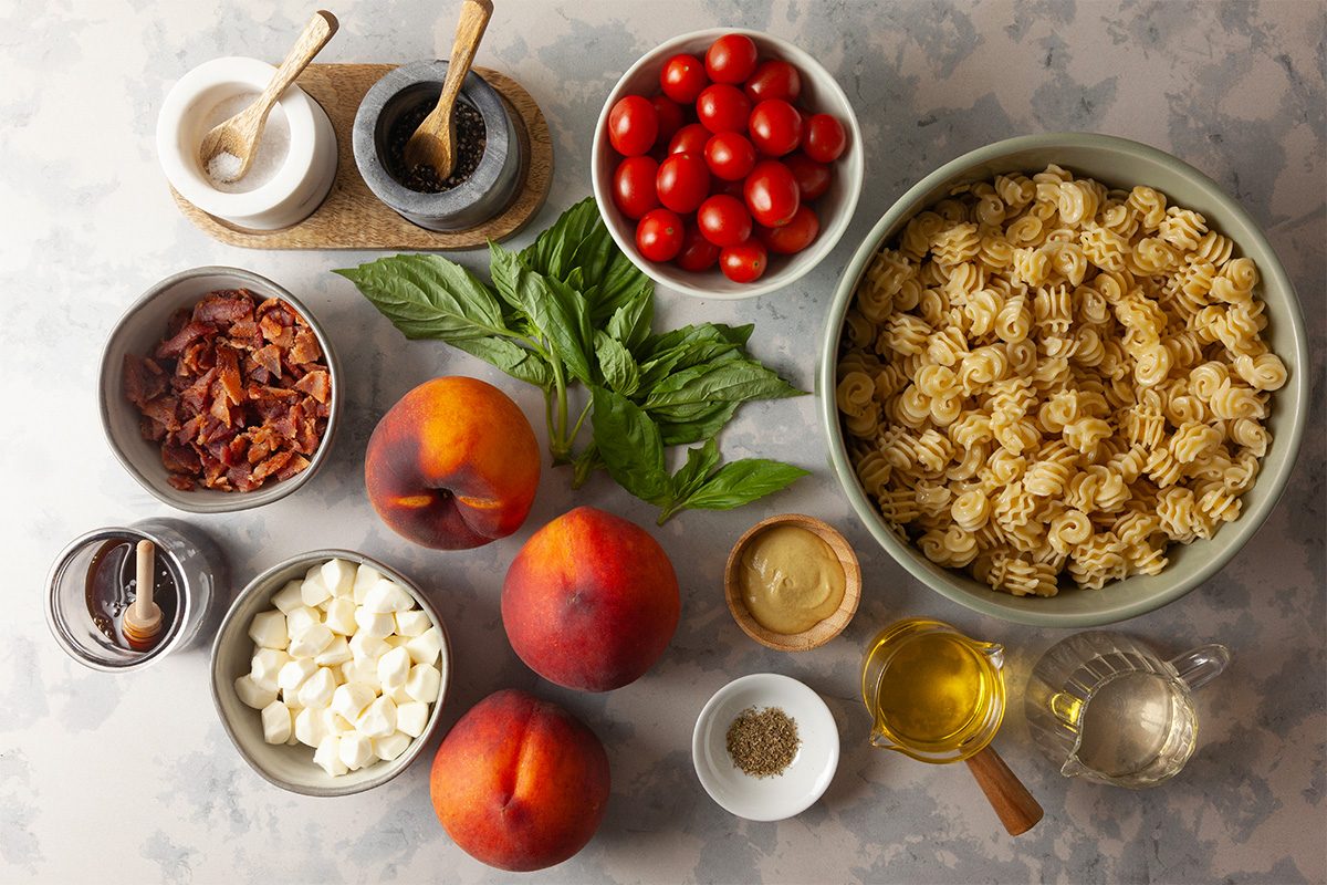 A flat lay of ingredients including cooked pasta, cherry tomatoes, crispy bacon, goat cheese, peaches, fresh basil, olive oil, vinegar, ground pepper, mustard, and salt and pepper shakers on a light surface.