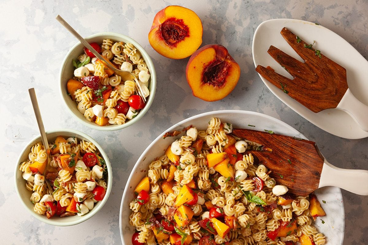 A large bowl and two smaller bowls filled with pasta salad containing cherry tomatoes, mozzarella balls, and diced peaches. Two peach halves and wooden salad utensils are nearby on a light gray surface.