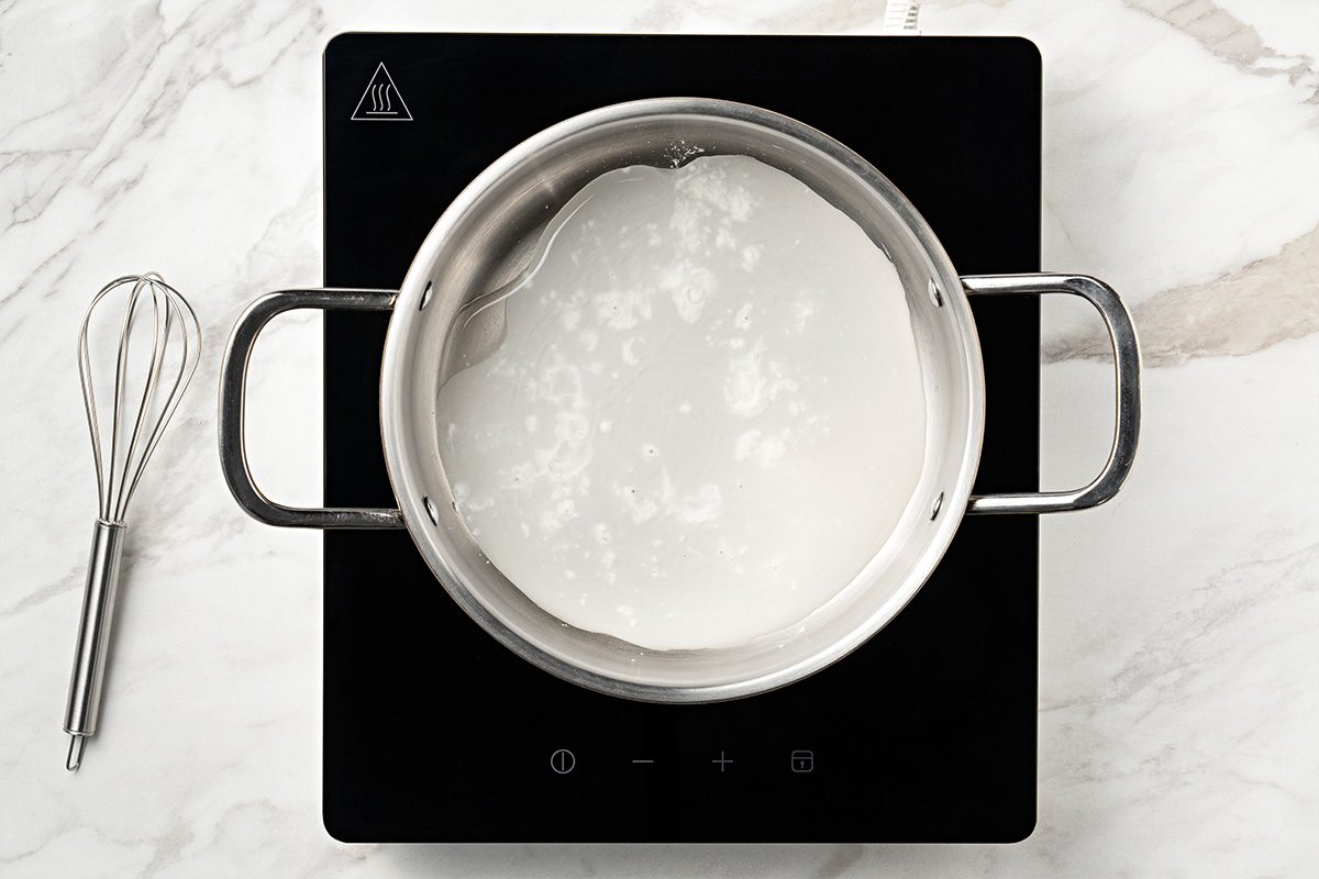 A stainless steel pot with milk is heating on a black induction cooktop, with a metal whisk placed nearby on a white marble countertop.
