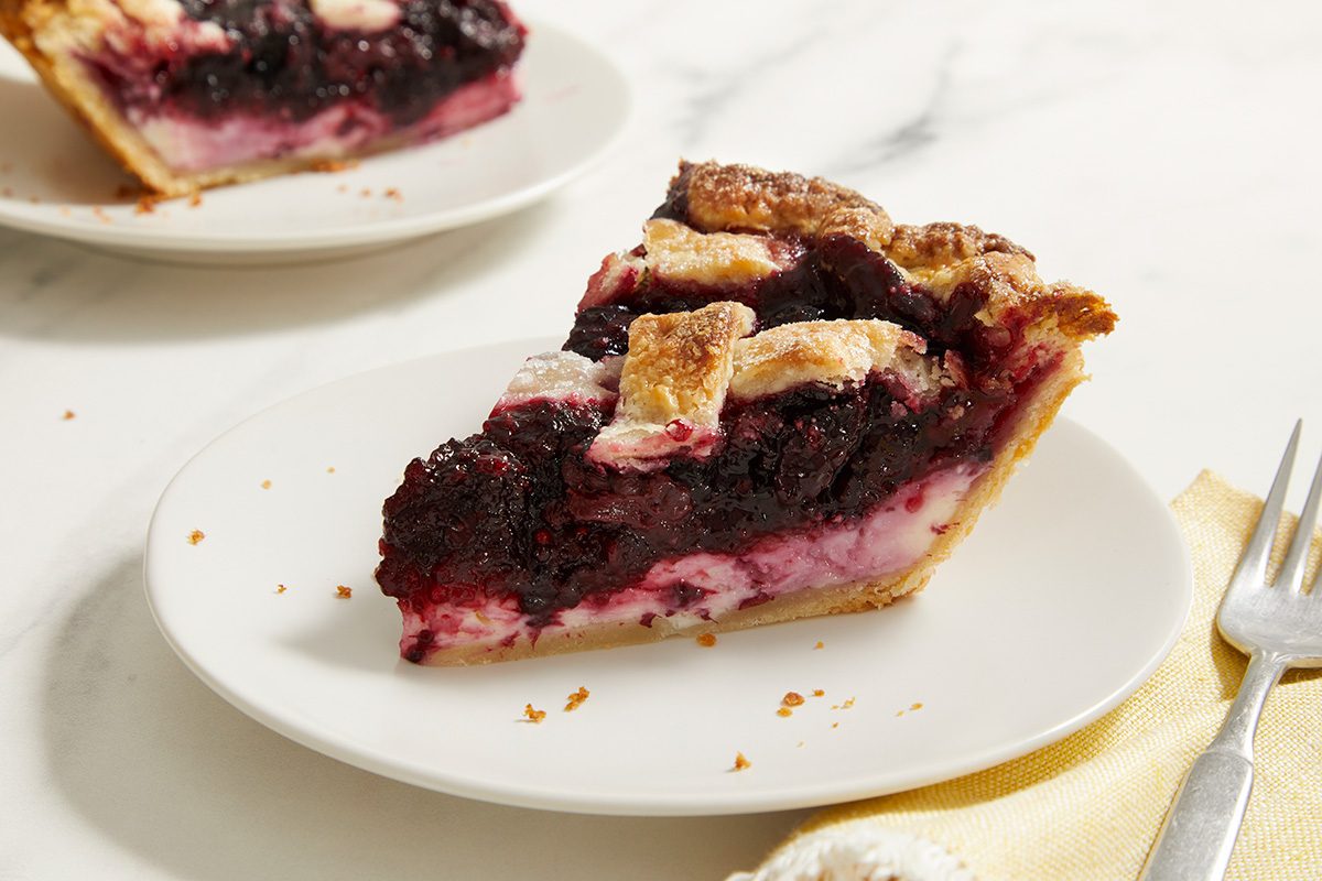 A slice of berry pie with a golden lattice crust sits on a white plate, showing layers of dark berry filling and creamy base. A fork and napkin are nearby, with another plate in the background.