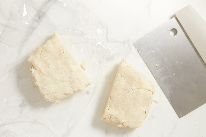Two pieces of dough sit on a sheet of plastic wrap next to a metal bench scraper on a white marble surface.