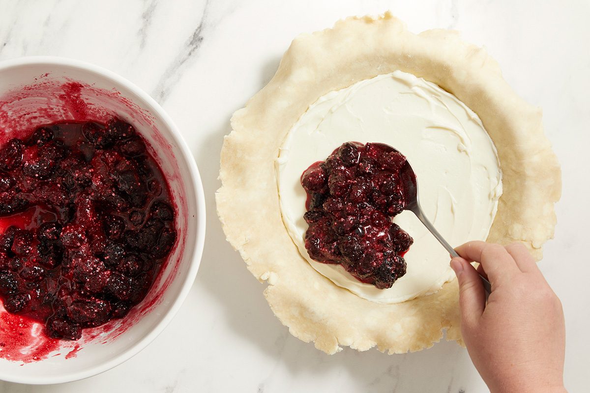 A hand spoons a berry filling from a bowl into a pie crust lined with a creamy white layer, on a white marble surface.
