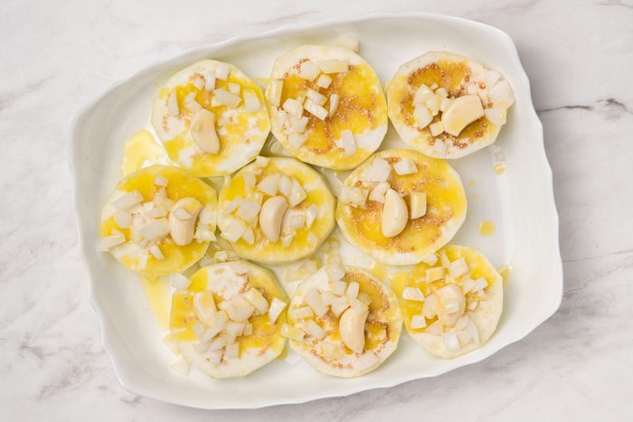 Overheda shot of a white dish with sliced eggplant topped with chopped onions, whole garlic cloves, and drizzled olive oil, set on a light marble surface;