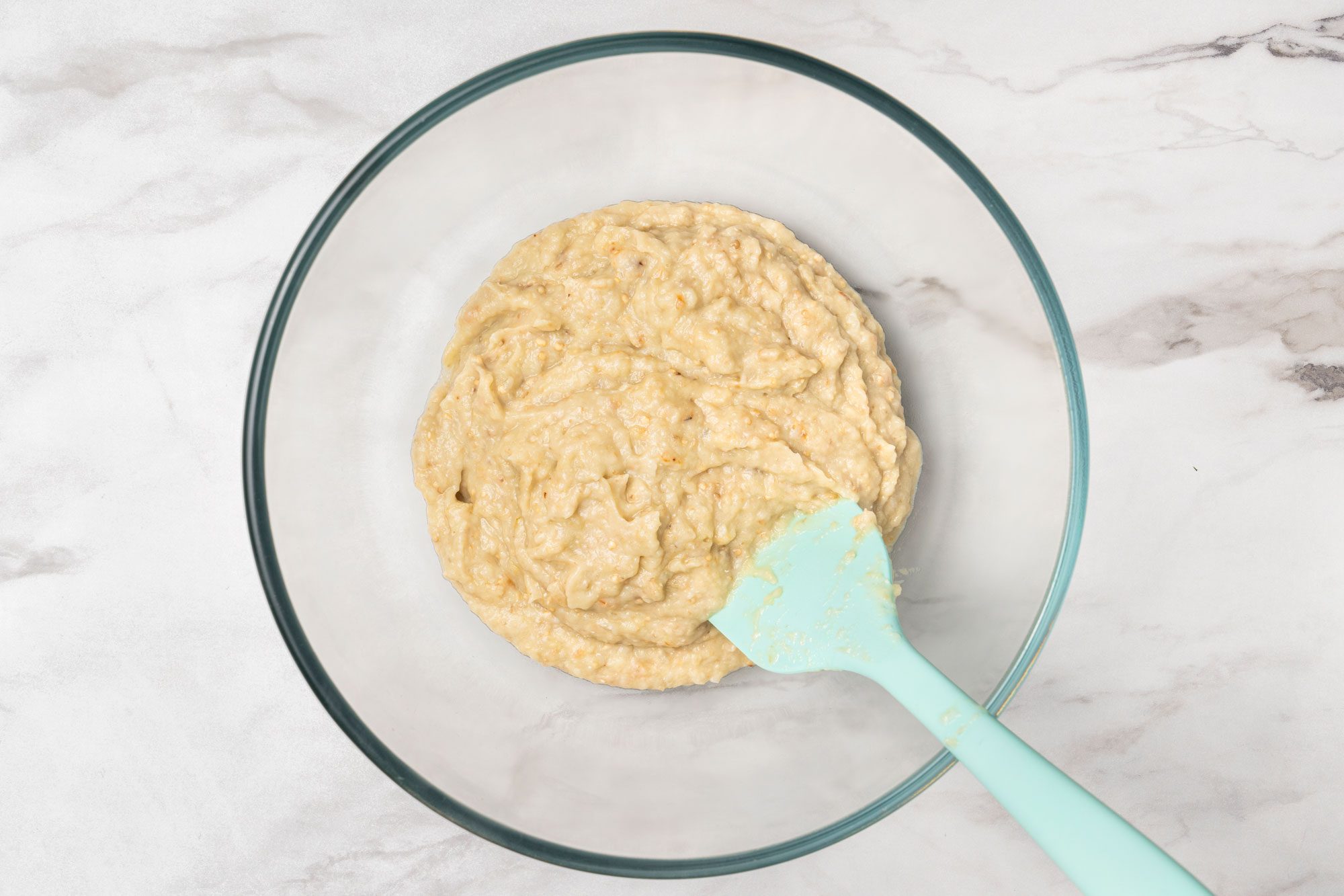 Overhead shot of a glass mixing bowl containing a thick, beige batter sits on a light marble surface; A light blue silicone spatula rests in the batter inside the bowl;