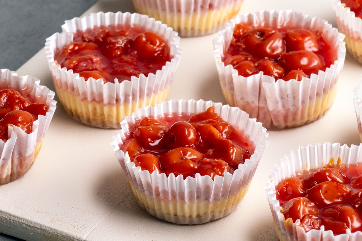 3/4th shot of Mini cheesecakes in white paper liners, topped with bright red cherry pie filling, are arranged on a light colored surface