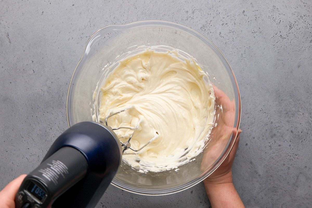 overhead shot of a hand holds a glass bowl filled with creamy batter while using a black electric hand mixer to blend the mixture on a gray countertop