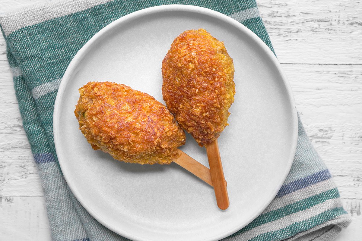 Two breaded, golden-brown chicken drumsticks on wooden sticks are served on a white plate, which sits on a green and white striped cloth, atop a white wooden surface.