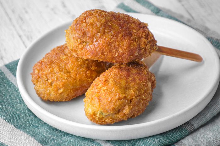 Three golden-brown, breaded corn dogs are served on a white plate, with one on a wooden stick, placed on a green and white striped cloth.