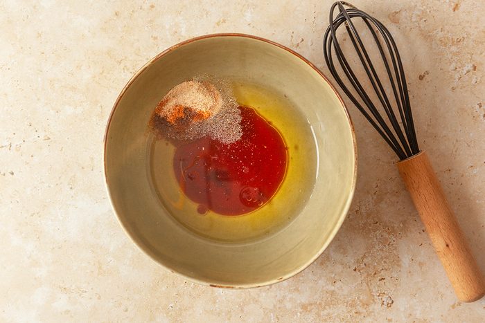 overhead shot of a green bowl containing oil, vinegar, and spices sits on a tan countertop next to a metal whisk with a wooden handle