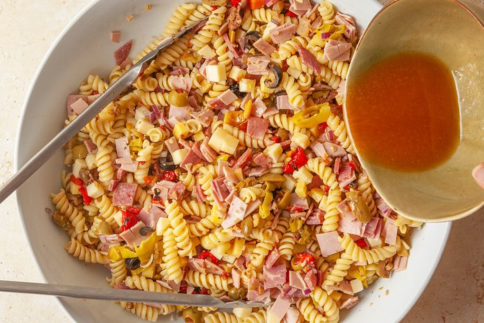 overhead shot of a large bowl of rotini pasta salad with chopped ham, cheese, olives, and peppers, with tongs on the side; A hand is pouring brown dressing from a small bowl onto the salad