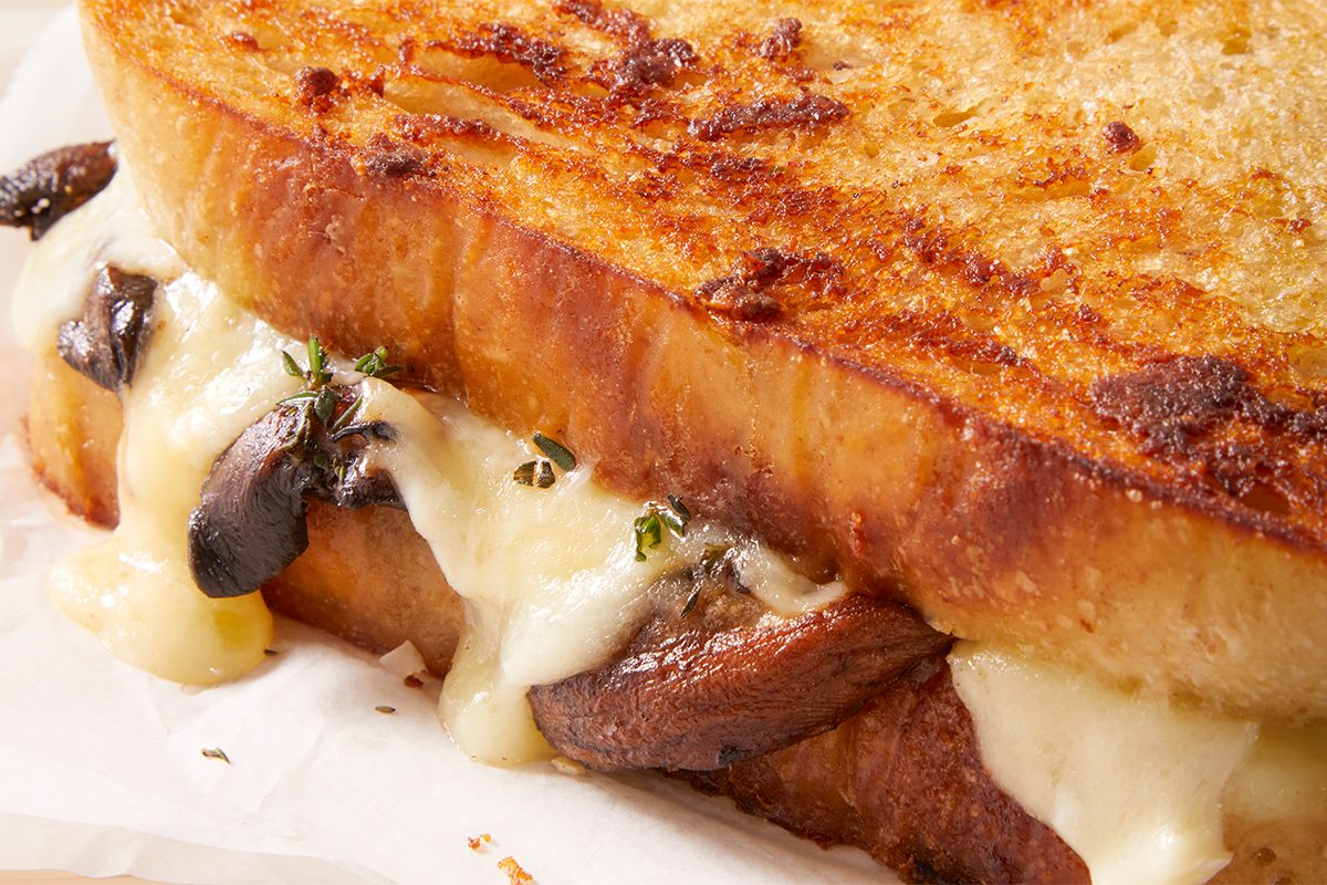 Close-up of a grilled cheese sandwich with melted cheese and sautéed mushrooms between thick slices of golden, crispy bread, garnished with a hint of fresh herbs.