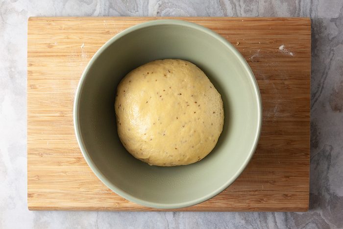 A ball of dough rests in a light green bowl placed on a wooden cutting board, viewed from above.