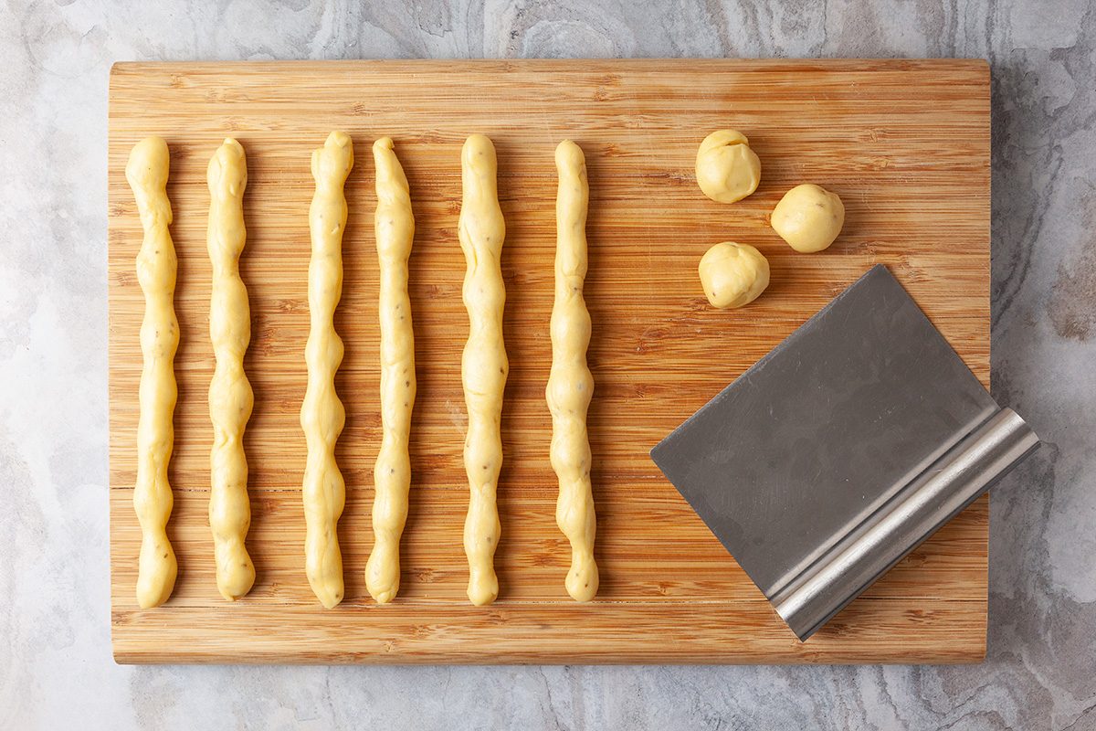 Six ridged dough logs, four dough balls, and a metal dough scraper are arranged on a wooden cutting board placed on a gray marble surface.
