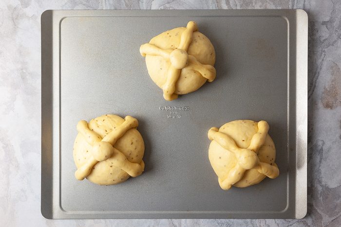 Three round pieces of dough with cross-shaped decorations on top are spaced apart on a silver baking sheet, ready to be baked.