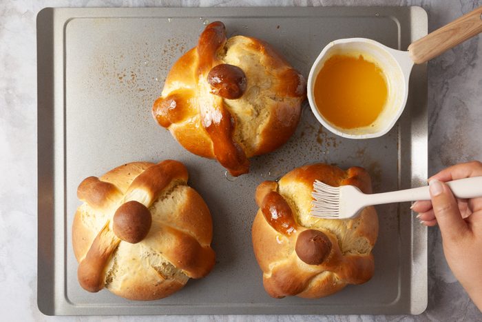 Three round loaves of bread with decorative dough shapes on top sit on a baking sheet. A hand uses a brush to glaze one loaf with a yellow liquid from a small white bowl.