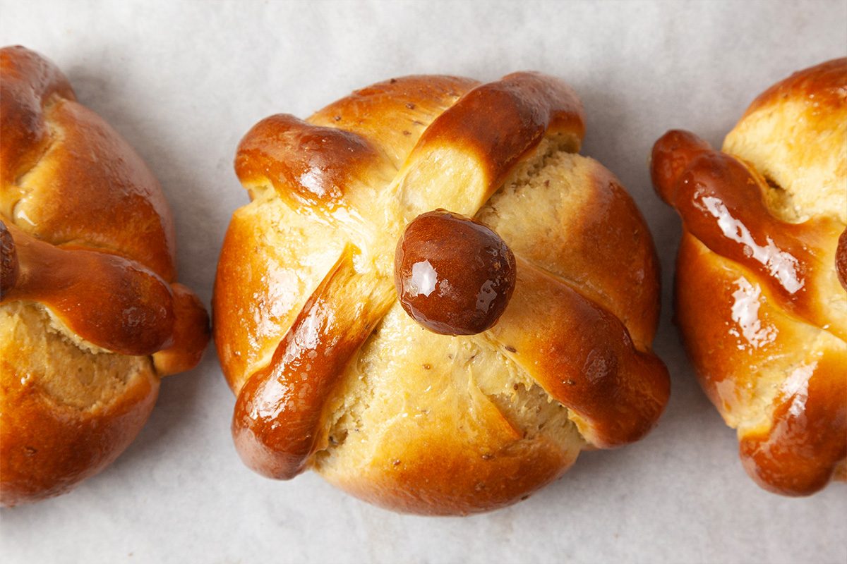 Three round pieces of pan de muerto bread, golden brown with shiny glaze, each decorated with dough shaped like bones arranged on top, placed on white parchment paper.