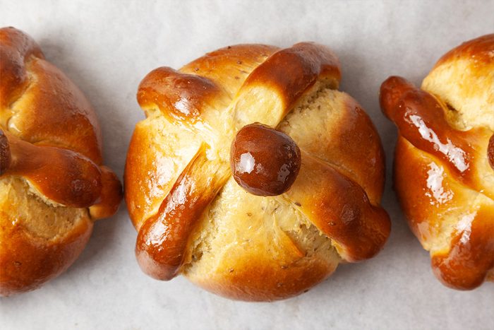 Three round pieces of pan de muerto bread, golden brown with shiny glaze, each decorated with dough shaped like bones arranged on top, placed on white parchment paper.