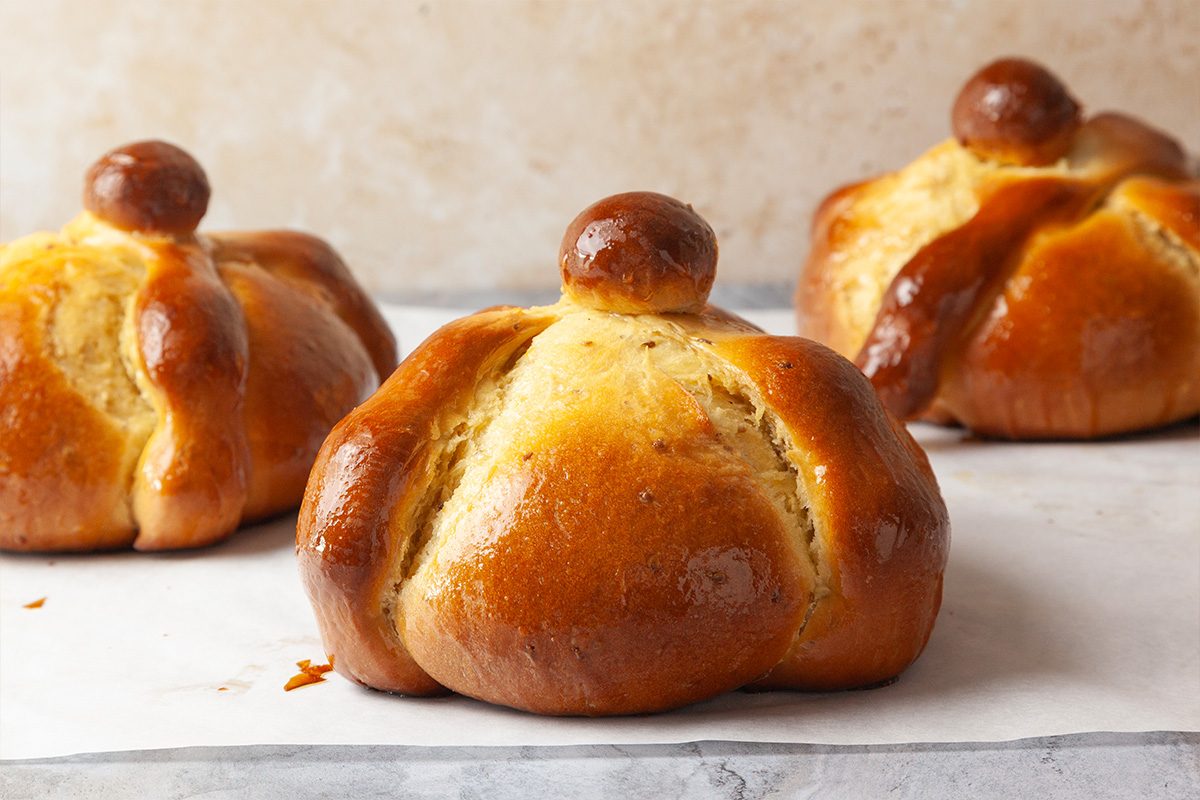 Three round, golden-brown pan de muerto breads with a glossy finish sit on a light surface. Each bread features a round knob on top and bone-shaped decorations across the top.