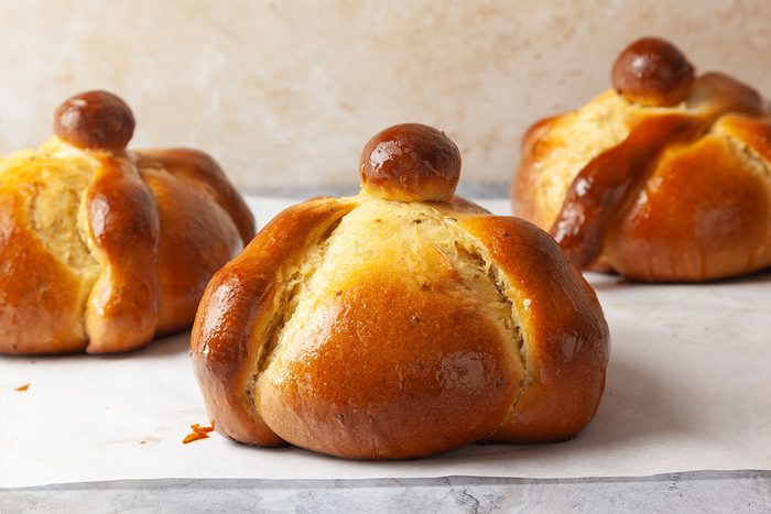 Three round, golden-brown pan de muerto breads with a glossy finish sit on a light surface. Each bread features a round knob on top and bone-shaped decorations across the top.
