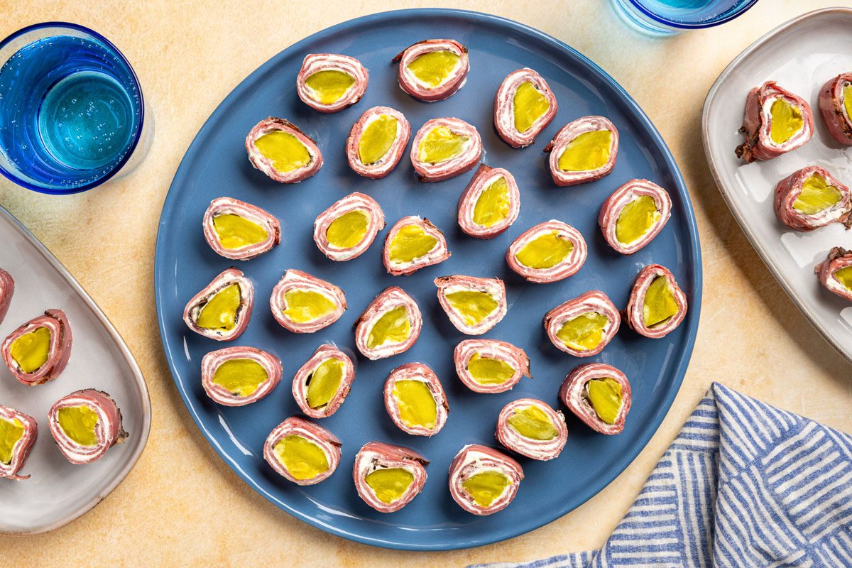 overhead shot of A blue plate holds neatly arranged Pastrami Roll Ups; Additional plates and blue glasses are nearby on a light table with a striped napkin