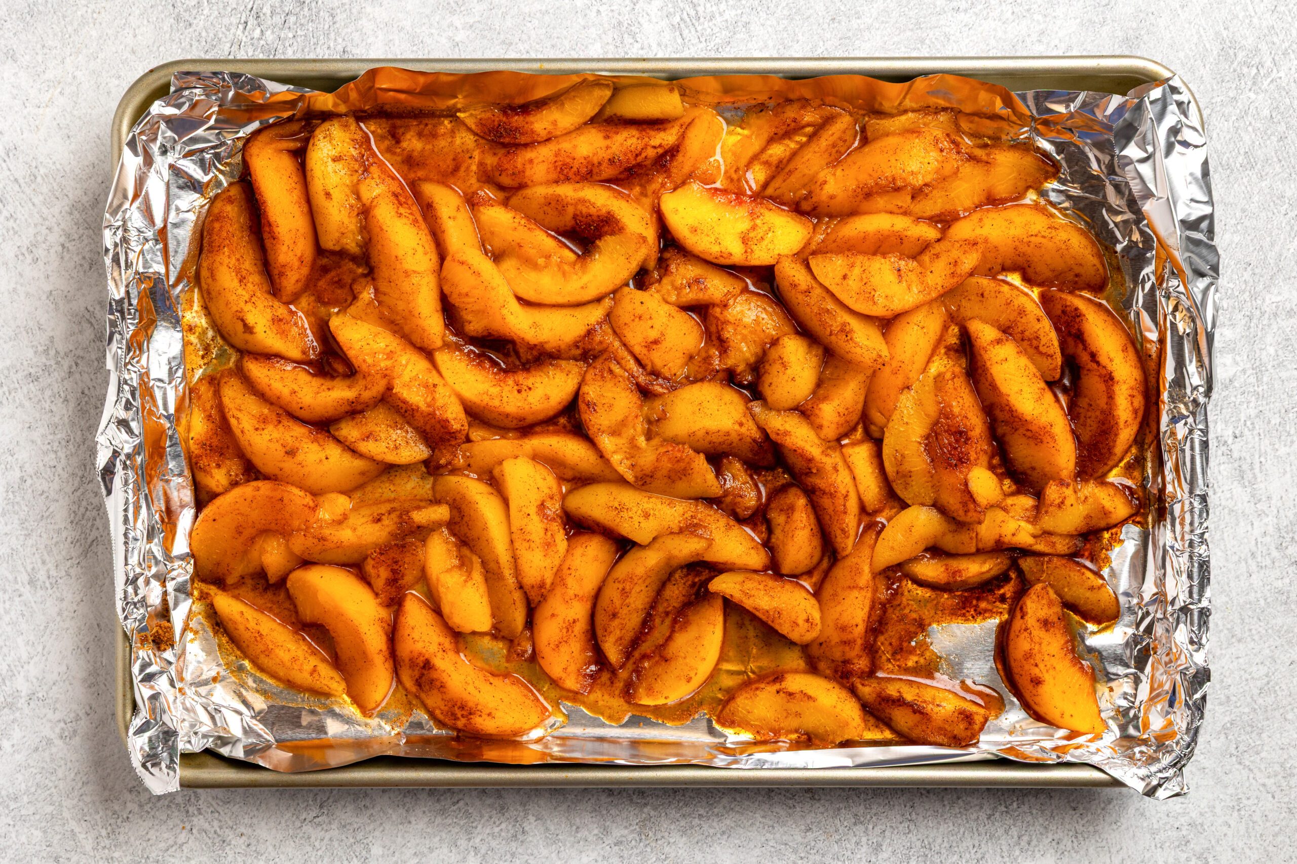 Peach slices coated with cinnamon and sugar are spread out on a foil-lined baking sheet, ready to be baked.