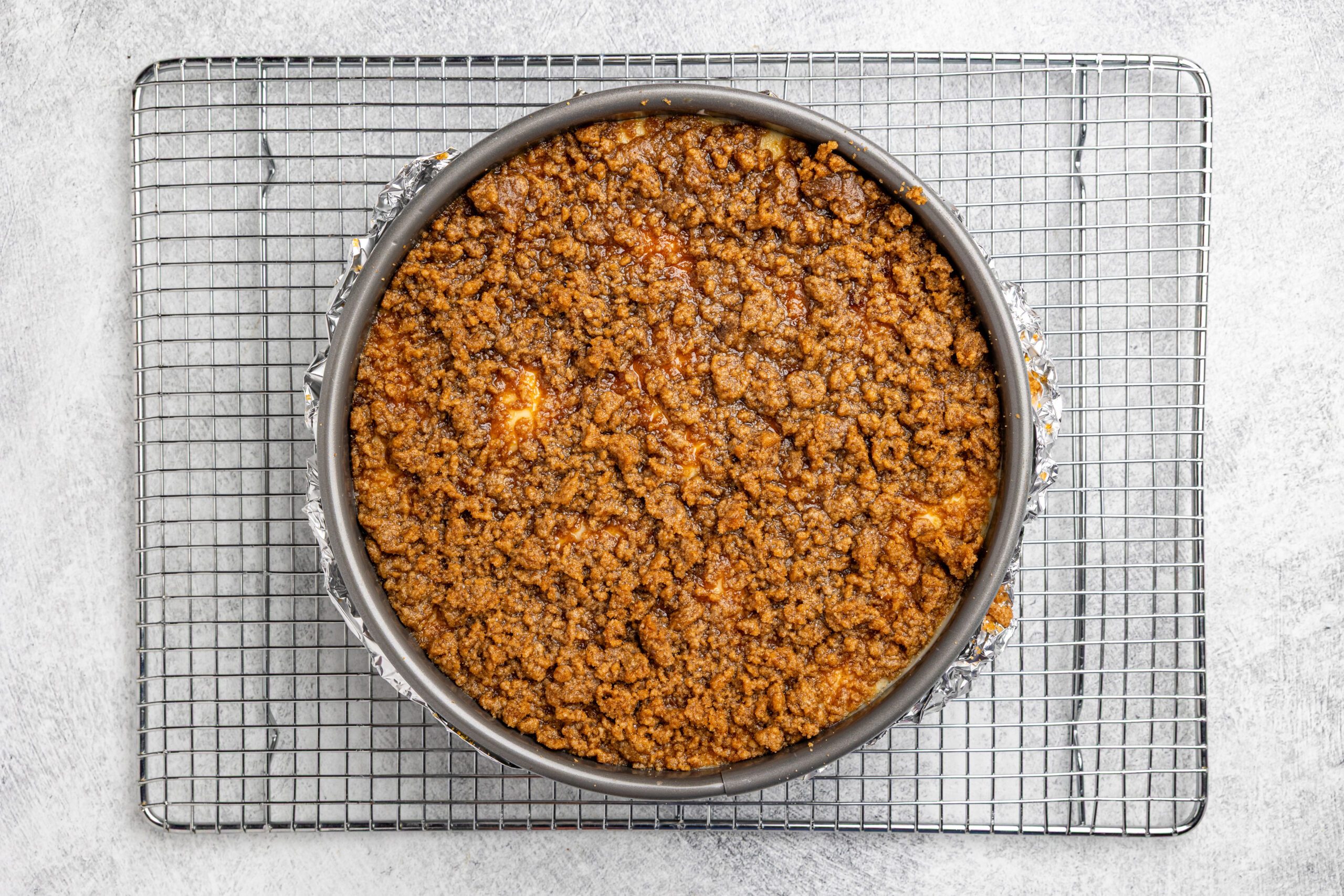 A round crumb cake with a golden-brown streusel topping sits in a baking pan on a wire cooling rack, viewed from above on a light gray surface.