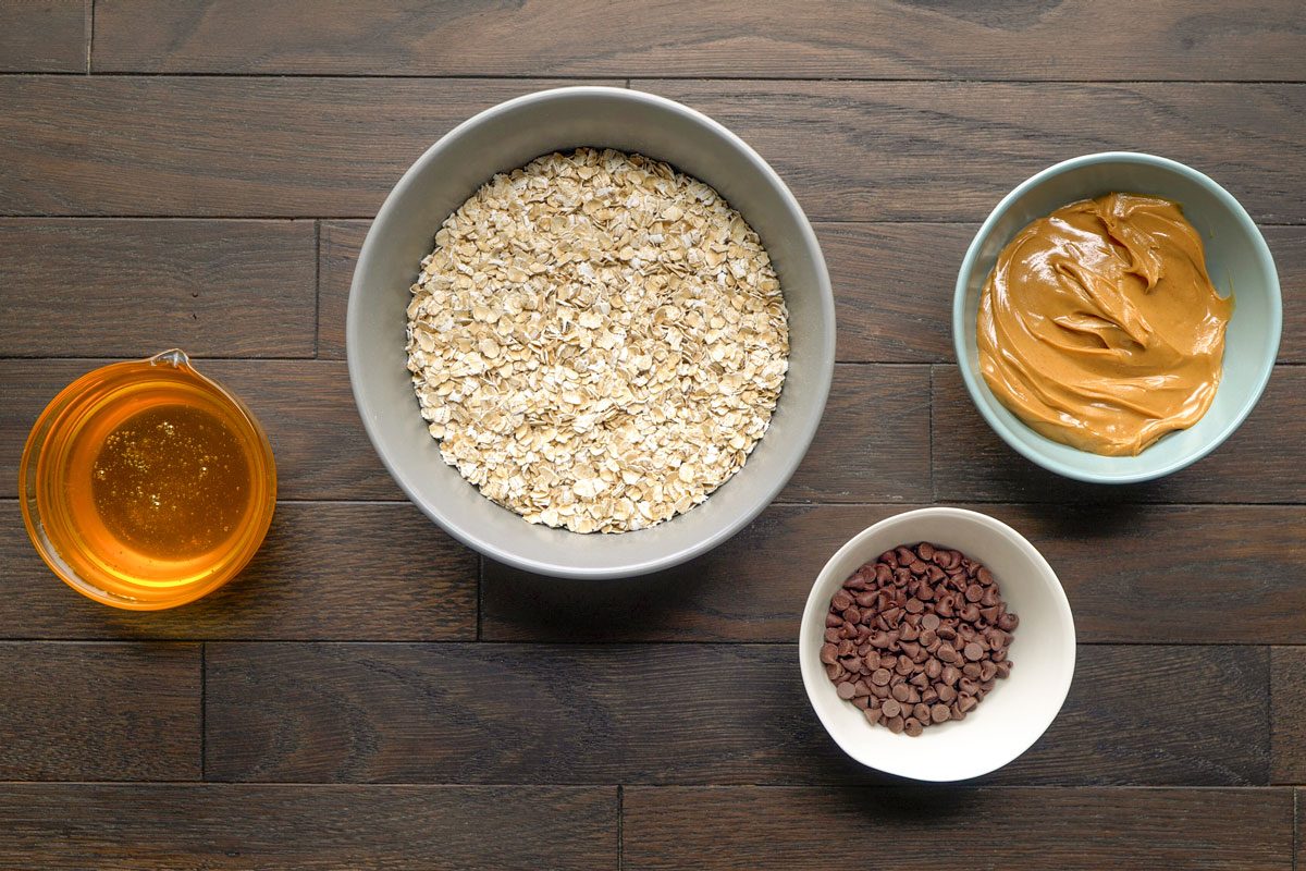 Overhead shot of four bowls on a wooden table; containing honey; rolled oats, creamy peanut butter; and mini chocolate chips;