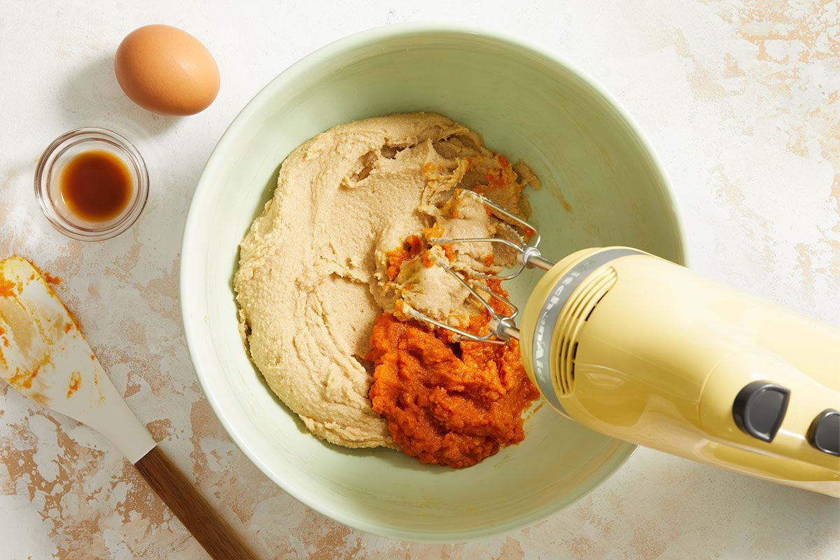 A yellow electric mixer blends brown sugar and pumpkin puree in a light green bowl. Nearby are a whole egg, a small bowl of vanilla extract, and a spatula with some mixture on it.