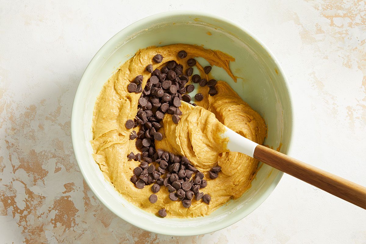 A mixing bowl containing cookie dough with chocolate chips on top and a spatula partially mixing the ingredients.