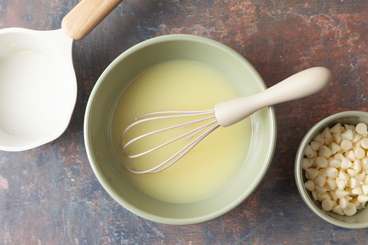 A whisk rests in a green bowl filled with melted yellow mixture. Nearby are a small pot of milk and a bowl of white chocolate chips, all on a textured brown surface.