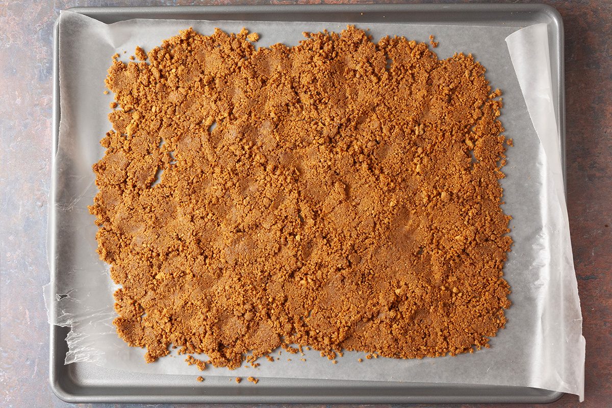 A baking sheet lined with parchment paper holds an even layer of crumbled brown mixture, likely crushed cookies or graham crackers, spread out and ready for baking.