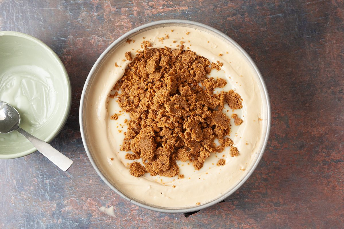 A round pan filled with creamy dessert topped with crumbled brown cookie or biscuit, next to a green bowl with a spoon on a rustic brown surface.