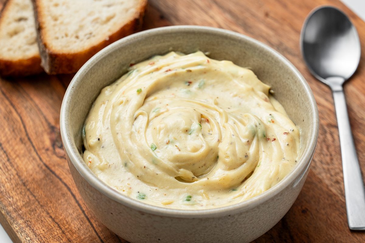 A bowl of creamy, herbed butter sits on a wooden board next to slices of bread and a spoon.