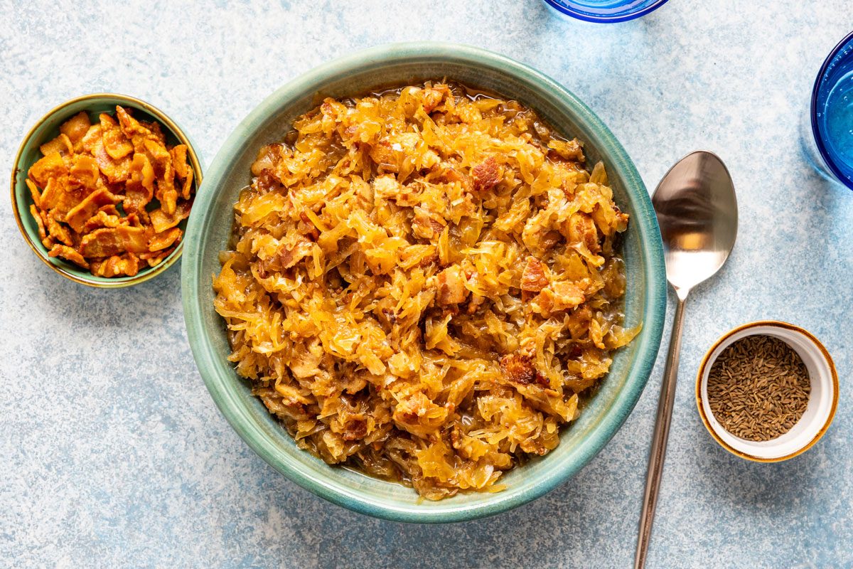 overhead shot of A bowl filled with cooked sauerkraut and sausage sits on a light surface, surrounded by a small dish of crispy toppings, a small bowl of caraway seeds, a spoon, and two blue glasses