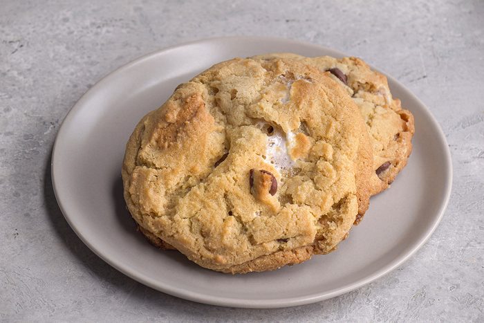 Two large chocolate chip cookies are stacked on a light gray plate, placed on a textured gray surface. The cookies look soft and slightly golden brown.