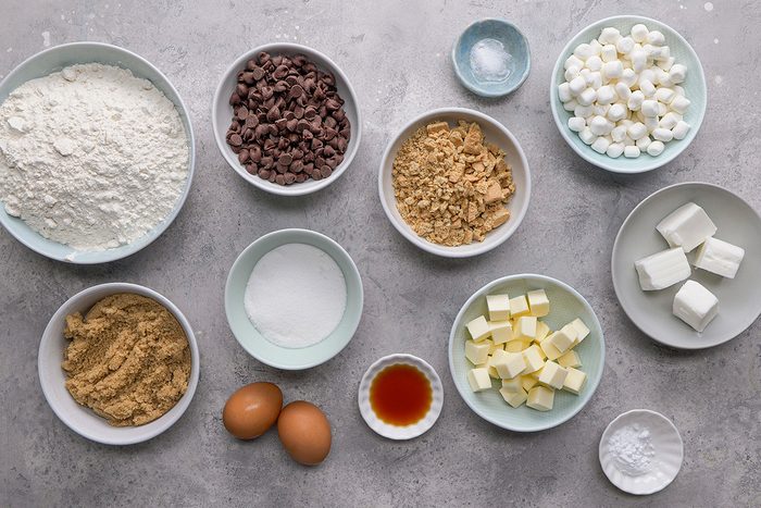 Various baking ingredients are arranged in bowls on a gray surface, including flour, chocolate chips, brown sugar, white sugar, mini marshmallows, eggs, butter cubes, vanilla extract, cream cheese, and baking powder.