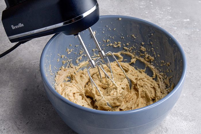 A hand mixer is blending a thick, beige batter in a large light blue mixing bowl on a gray countertop.