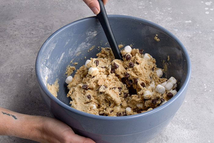A person stirs cookie dough with chocolate chips and mini marshmallows in a large blue mixing bowl using a black spatula.