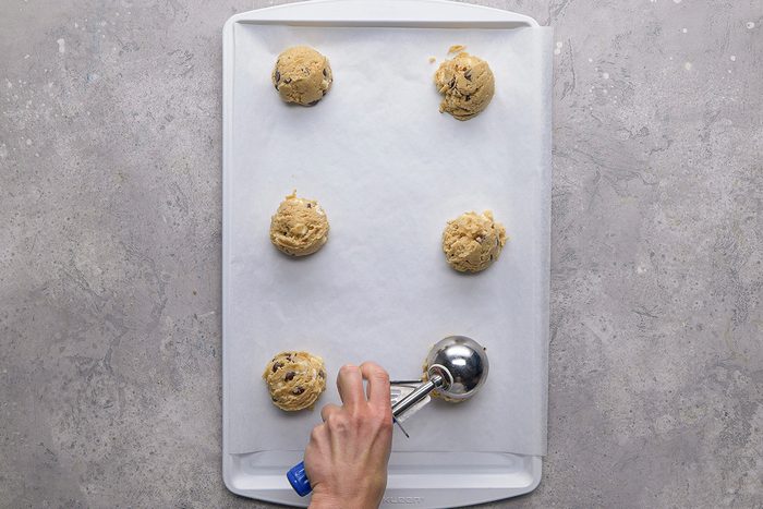 A hand uses a cookie scoop to place cookie dough balls on a baking sheet lined with parchment paper; five dough balls are already spaced on the sheet.