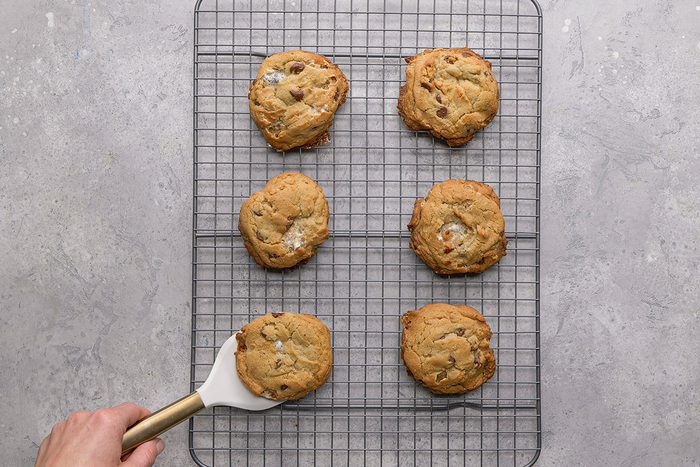 Six chocolate chip cookies are cooling on a metal wire rack over a gray surface. A hand using a spatula is about to lift one of the cookies from the rack.