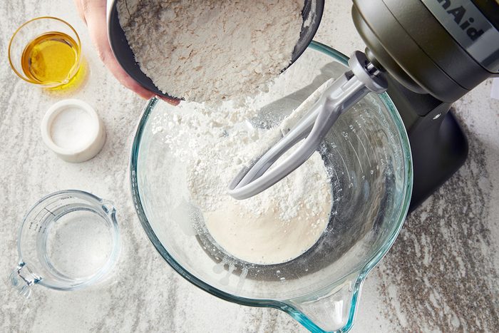 Overhead shot of a person pouring flour from a bowl into a stand mixer with dough; a glass of water, a bowl of salt; and a dish of oil are arranged on the kitchen counter nearby.