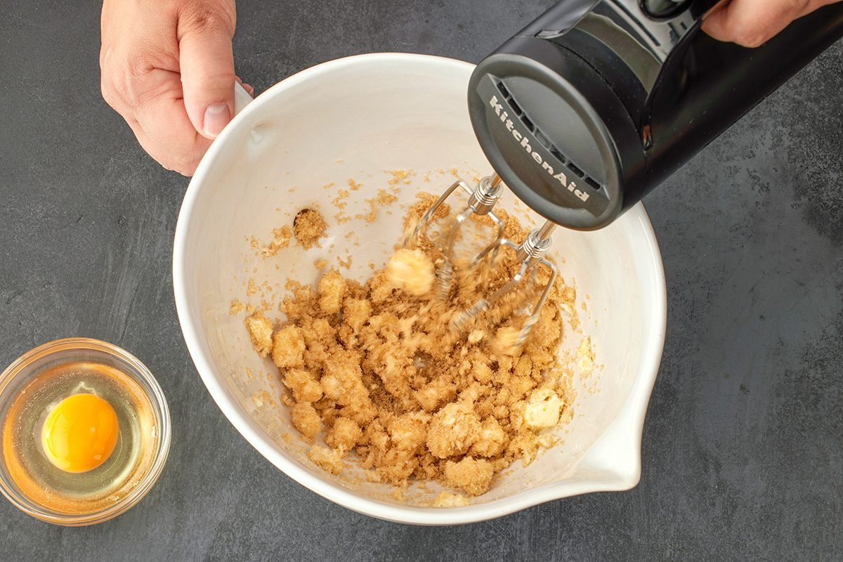 Overhead shot of a hand holding a white mixing bowl with sugar and butter being blended by a black electric mixer; while a small glass bowl containing a raw egg rests nearby on a dark countertop