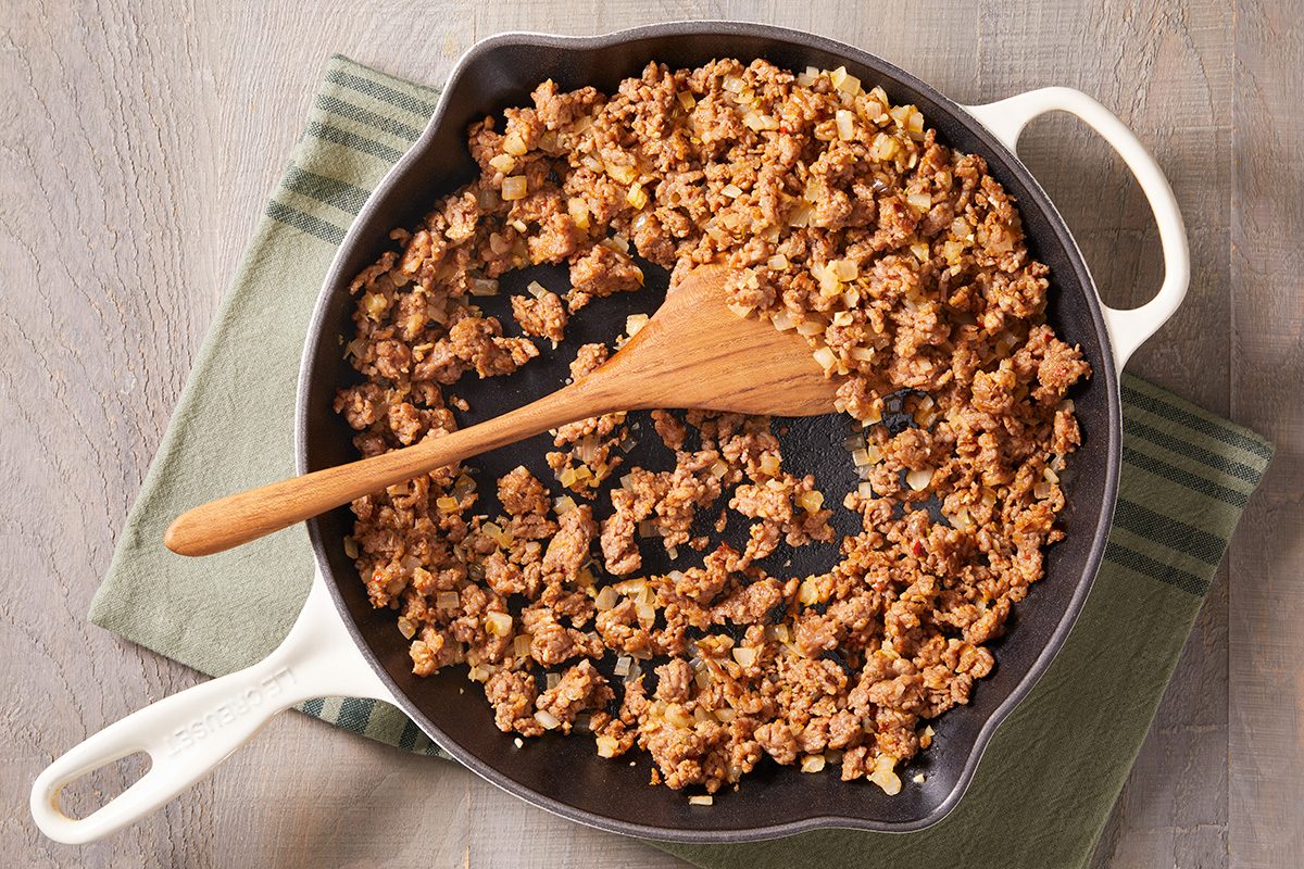 A white skillet on a green-striped towel contains cooked, crumbled ground meat being stirred with a wooden spatula.