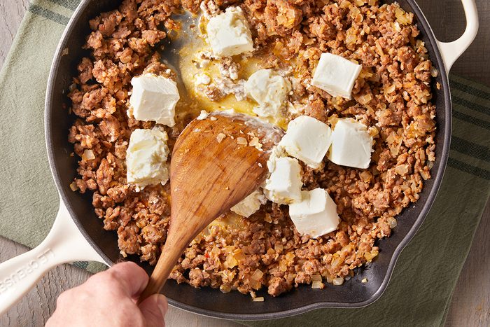 A hand stirs ground meat and onions in a skillet with several cubes of cream cheese melting on top.
