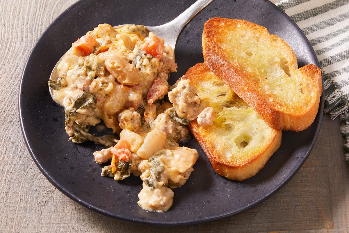 A dark plate with a serving of seafood casserole and two slices of toasted bread. A spoon is resting on the plate, and part of a striped cloth napkin is visible in the background.