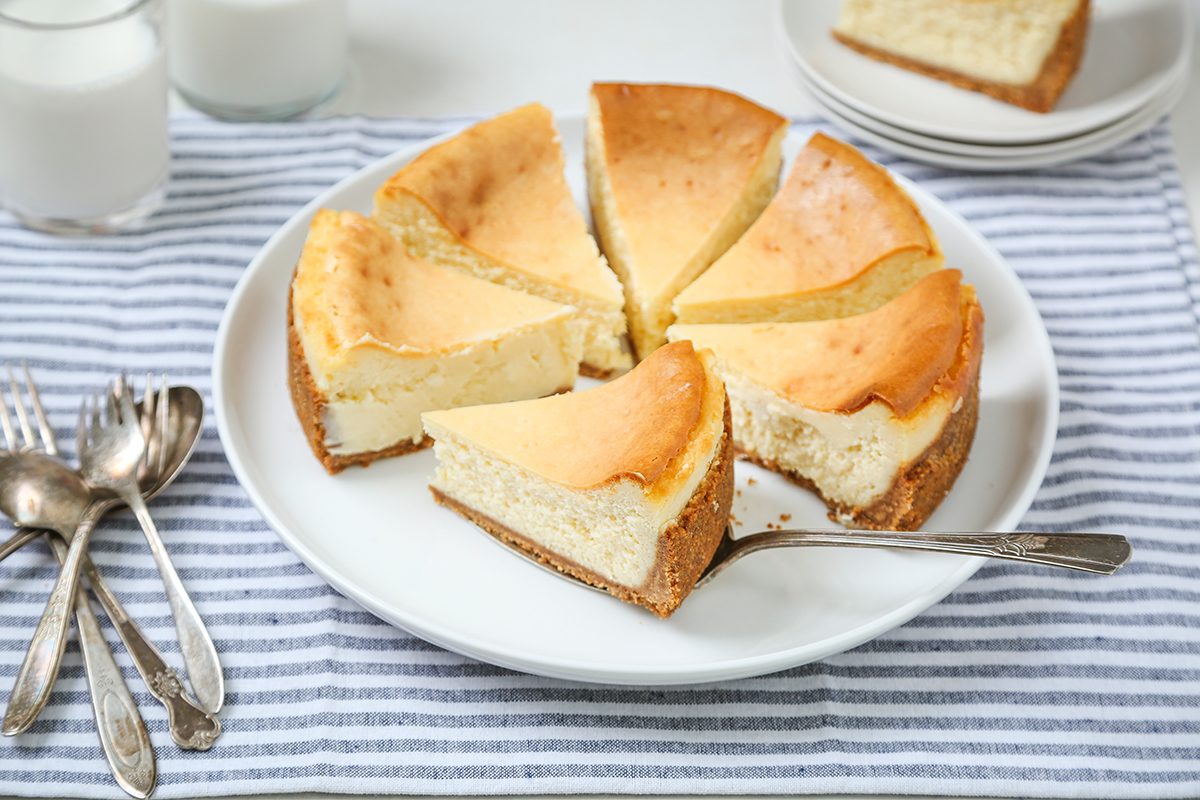 A round cheesecake cut into six slices sits on a white plate with a silver spoon, resting on a striped cloth. Nearby are glasses of milk, stacked plates, and a few spoons.