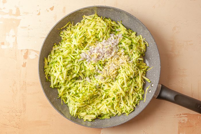 This is a close-up top-down shot; a frying pan filled with shredded zucchini; finely chopped onions; and minced garlic on a light background; ingredients appear fresh and uncooked