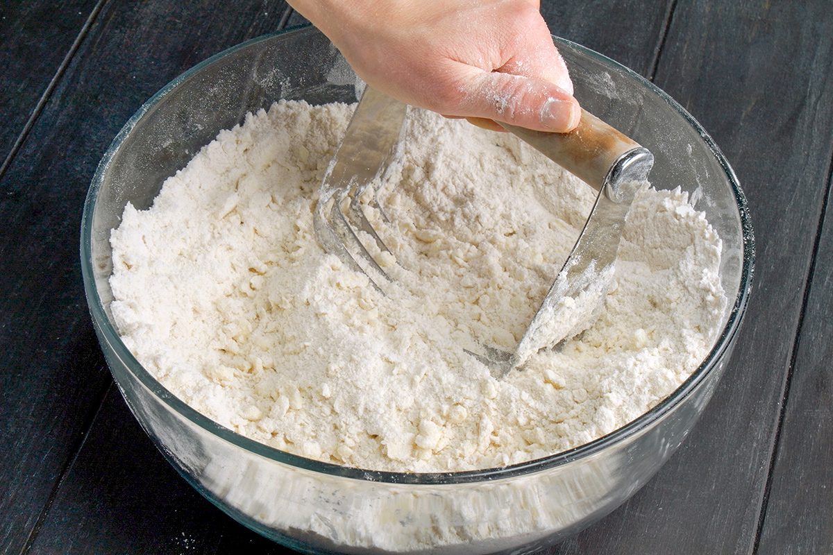 A hand uses a pastry blender to mix flour in a glass bowl on a dark wooden surface.